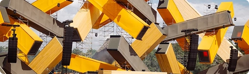Openluchtmuziekfestival met een grote menigte voor een podium met torenhoge abstracte gele en grijze geometrische structuren onder een gedeeltelijk bewolkte hemel