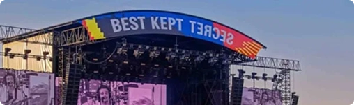 Openluchtmuziekfestival met een groot podium met het opschrift ‘Best Kept Secret’. Een band treedt op terwijl een enorme menigte geniet van de show onder een strakblauwe hemel