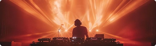 DJ treedt op op het podium, omringd door levendige oranje en rode lichten die dynamische stralen creëren. De sfeer is energiek en meeslepend