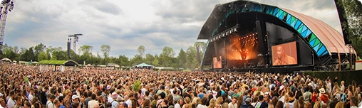 Een groot publiek op een openluchtmuziekfestival onder een bewolkte hemel, met uitzicht op een helder verlicht podium. De sfeer is levendig en druk, en er hangt een opgewonden stemming in de lucht