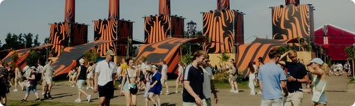 Buitenfestival met mensen die rondlopen en kletsen. Levendige oranje en zwart gestreepte torens domineren de achtergrond. De sfeer is levendig en ontspannen