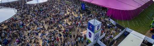 Luchtfoto van een groot muziekfestival met mensenmassa's verzameld voor een opvallende paarse tent. De lucht is gedeeltelijk bewolkt en de sfeer is levendig