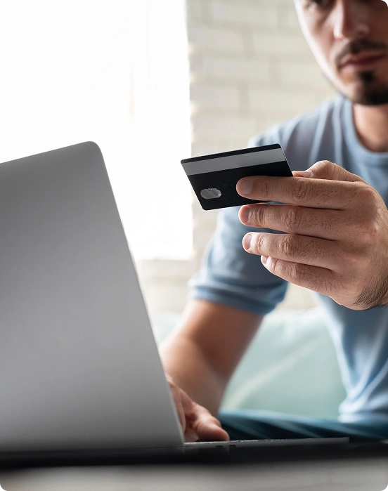 Een man in een blauw shirt die een laptop gebruikt terwijl hij een creditcard vasthoudt, wat duidt op online winkelen of bankieren. De omgeving is helder en scherp in beeld