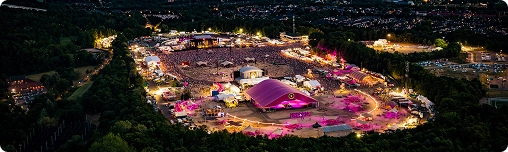 Luchtfoto van een levendig muziekfestival bij zonsondergang, met kleurrijke lichten die tenten en podia verlichten. Een grote menigte verzamelt zich onder een dramatische avondhemel