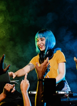 DJ met blauw haar en koptelefoon lacht terwijl hij muziek mixt op een laptop. Enthousiast publiek steekt de handen in de lucht in een rokerige, kleurrijke sfeer. Opgewekte stemming