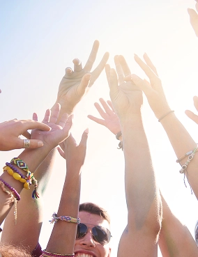 Handen van diverse mensen, versierd met kleurrijke armbanden, reiken omhoog naar de zonnige hemel. Het tafereel straalt vreugde en eenheid uit